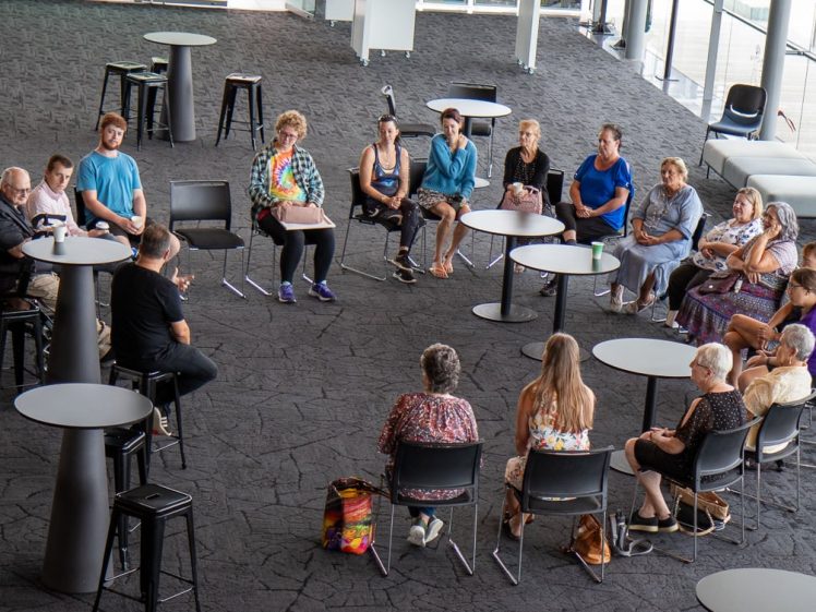 Image of A group of people sit in a large circle on chairs in a modern, open room with round tables and carpeted floor, engaged in a discussion or meeting.