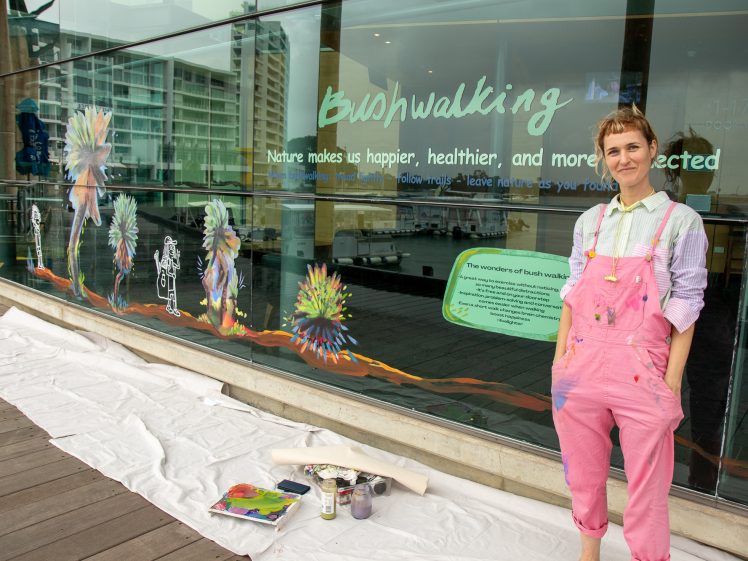 Image of A person in pink dungarees and a striped shirt stands beside a glass wall decorated with colourful art and the word Bushwalking, with art supplies and a white sheet on the ground.