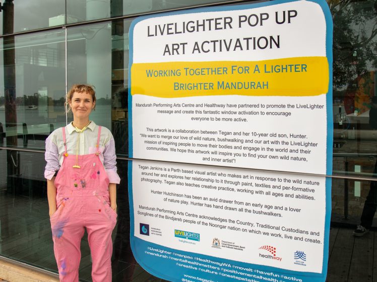 Image of A person in pink dungarees and a striped shirt stands smiling beside a large sign for the LiveLighter Pop Up Art Activation outside a building, with details about the collaborative art event in Mandurah.
