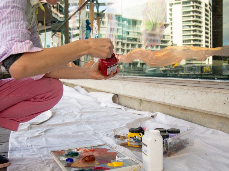 Image of A woman kneels on a white sheet, painting a mural on a glass wall outdoors. Paints, brushes, and art supplies are spread out beside her. City buildings are visible in the background.