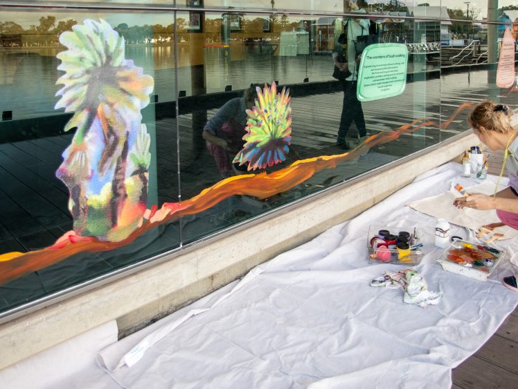Image of A person sits on a white sheet painting colourful, abstract designs on a large glass window by the waterfront. Art supplies and paint jars are spread on the sheet next to them.