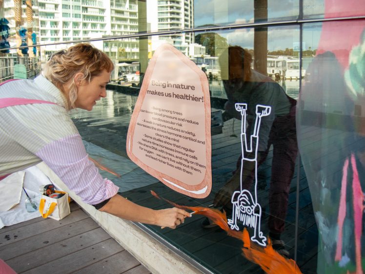 Image of A woman in dungarees paints an orange design on a window next to a white drawing of a person doing a handstand and a sign about nature’s health benefits. Buildings, trees, and water are visible in the background.