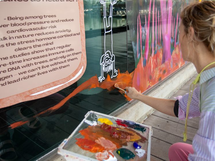 Image of A person with a paint palette paints an orange landscape on a glass window, where white line art of a figure and text about trees and health benefits are also visible.
