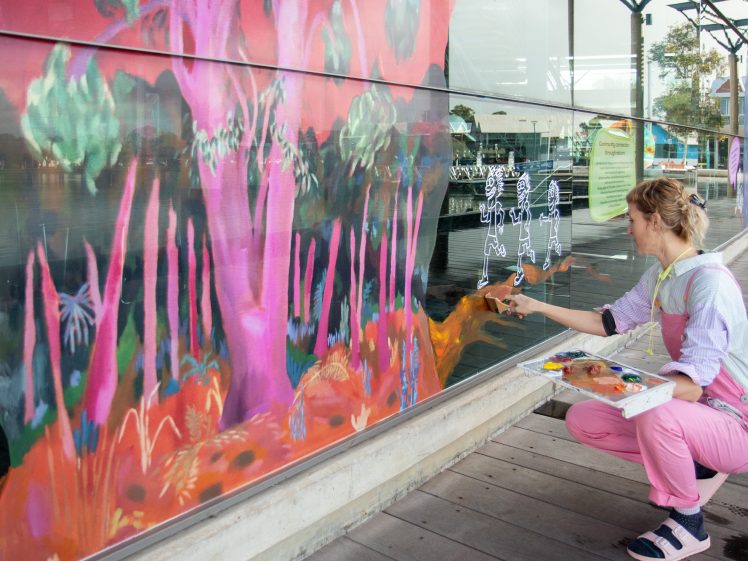 Image of A person in pink dungarees paints vibrant, colourful trees and white outlined figures on a large glass window outdoors, holding a palette and brush near a waterfront.
