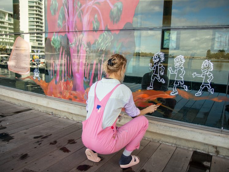 Image of A person in pink dungarees is crouched down, drawing white chalk figures of running children on a window decorated with a colourful mural. Modern buildings and water are visible in the reflection.