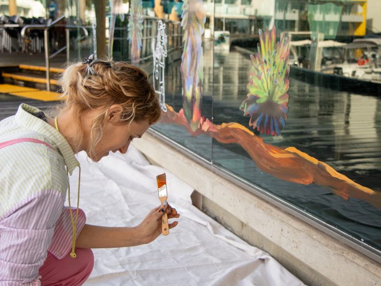 Image of A woman kneels on a white sheet, painting a colourful mural on a glass wall outdoors. She holds a paintbrush and palette, with various paints and supplies beside her. Buildings and reflections are visible in the background.