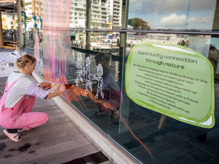 Image of A woman in pink dungarees paints white cartoon figures on a glass wall next to a large sign promoting community nature activities, beside a marina with boats in the background.
