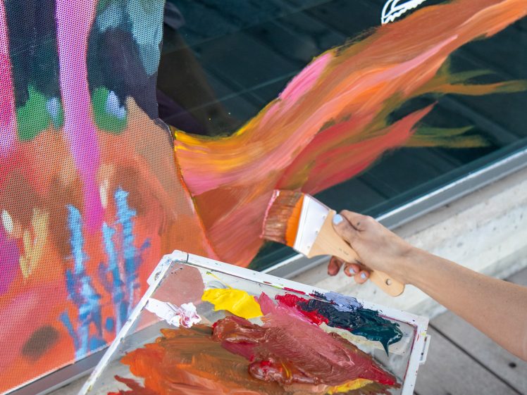 Image of A person holding a palette with colourful paint uses a brush to paint vibrant orange and pink strokes on a glass window, which also has a white drawing of a running figure.