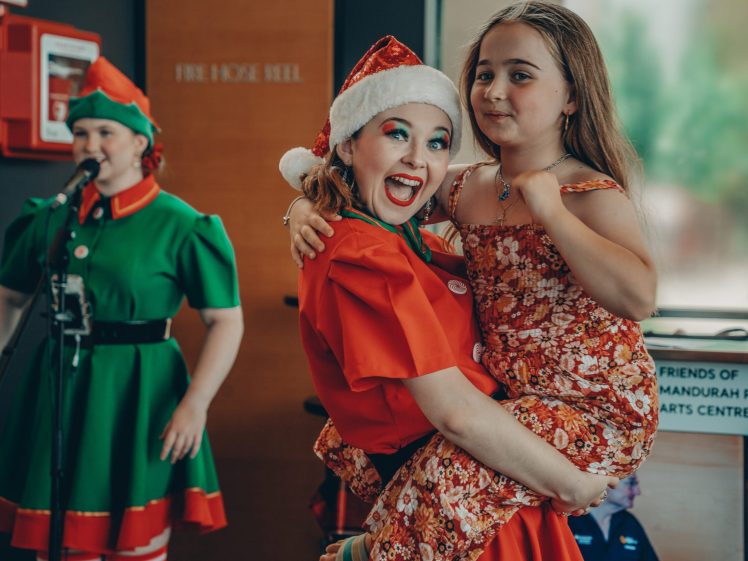 Image of A smiling woman in a Santa hat and festive clothes holds a young girl in a red floral dress. Another woman in an elf costume sings into a microphone in the background. The scene is cheerful and indoors.