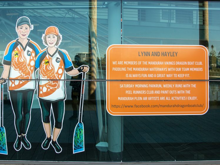 Image of Two illustrated women in orange uniforms holding paddles stand next to a sign about Lynn and Hayley, members of the Mandurah Vikings Dragon Boat Club, with club activities listed and a Facebook link on a glass wall.