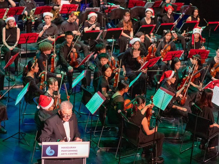 Image of A large orchestra, many musicians wearing Santa hats, performs on stage. A man stands at a lectern labelled Mandurah Performing Arts Centre in front of the ensemble, suggesting a festive or holiday concert.