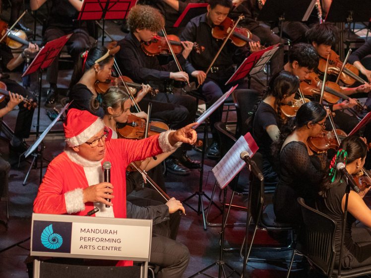 Image of A person dressed as Father Christmas, holding a microphone and pointing, stands at a lectern labelled Mandurah Performing Arts Centre, in front of an orchestra playing string instruments.