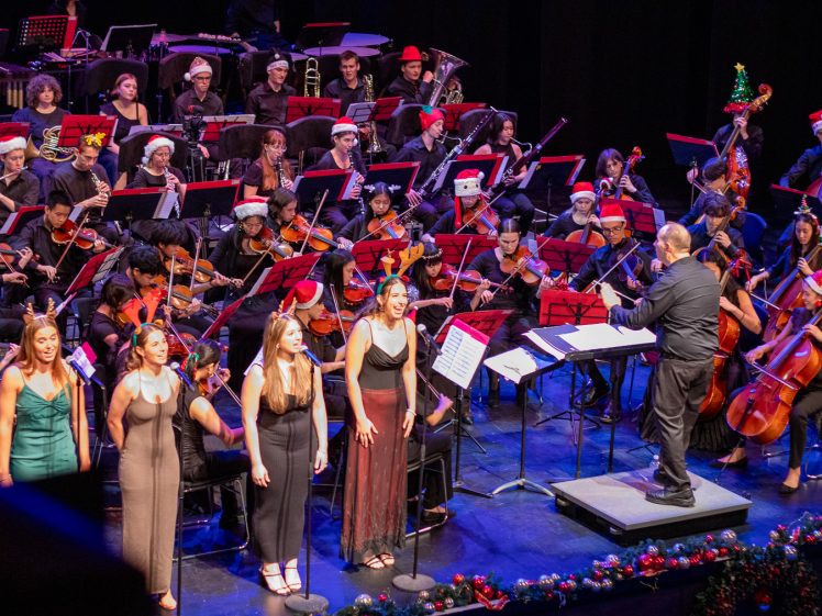Image of A large orchestra wearing festive hats performs on stage whilst a conductor leads. Four women in formal dresses stand at microphones singing in front of the musicians. Holiday decorations line the stage.