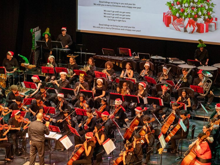 Image of A symphony orchestra in festive attire performs on stage with a decorated Christmas tree and carol lyrics projected behind them. Many musicians wear Father Christmas hats and the stage is adorned with holiday decorations.