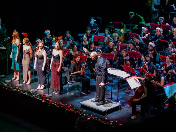 Image of A conductor bows before an orchestra on stage, with four female vocalists standing to the left in festive attire. Musicians wear holiday hats, and Christmas decorations line the front of the stage.