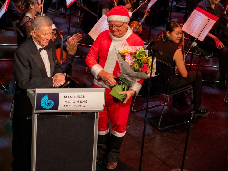 Image of A man in a black dinner jacket speaks at a podium labelled Mandurah Performing Arts Centre while another man dressed as Father Christmas holds flowers on stage, with orchestra members seated behind them.