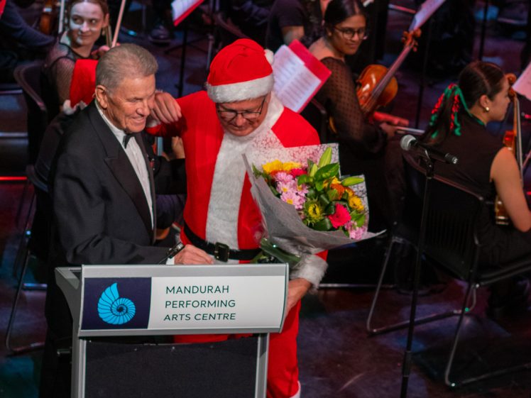 Image of An older man in a suit and a man dressed as Father Christmas holding flowers stand together at a podium labelled Mandurah Performing Arts Centre, with an orchestra seated behind them.