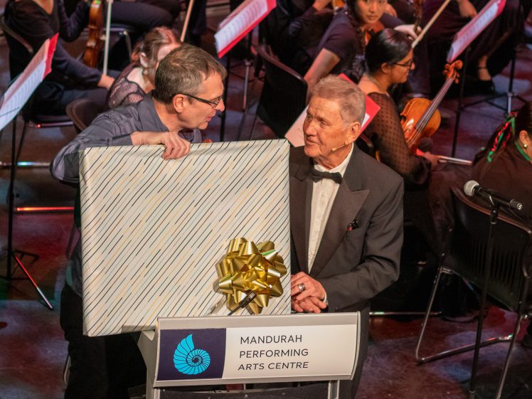 Image of Two men stand at a podium labelled Mandurah Performing Arts Centre. One holds a large, wrapped present with a gold bow. An orchestra is seated behind them, and both men are dressed formally.