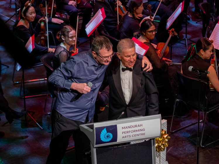 Image of Two men stand together smiling at a lectern labelled Mandurah Performing Arts Centre on stage, with an orchestra and musicians playing string instruments seated behind them. The lectern is decorated with a gold ribbon.