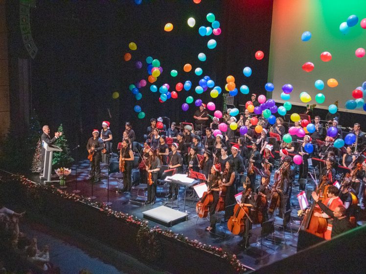 Image of A large orchestra on stage, some members wearing Father Christmas hats, is applauded by an audience as colourful balloons fall from above. The stage is decorated with festive lights and garlands, with a conductor standing at a lectern.