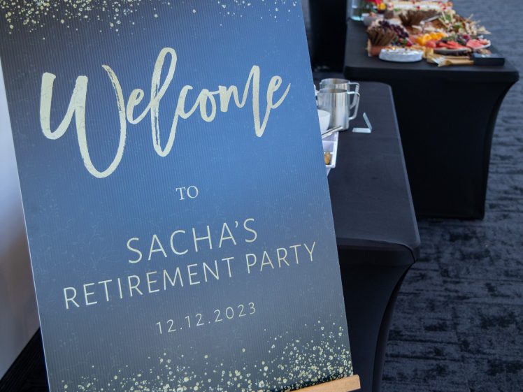 Image of A sign on an easel reads “Welcome to Sacha’s Retirement Party 12.12.2023” in gold and white lettering. A table with food and drinks is visible in the background.