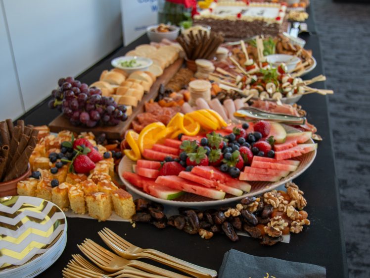 Image of A table set with a variety of fruits, cheeses, desserts, and snacks, alongside gold forks, patterned plates, and black serviettes with Happy Birthday written in gold. Flowers are seen in the background.