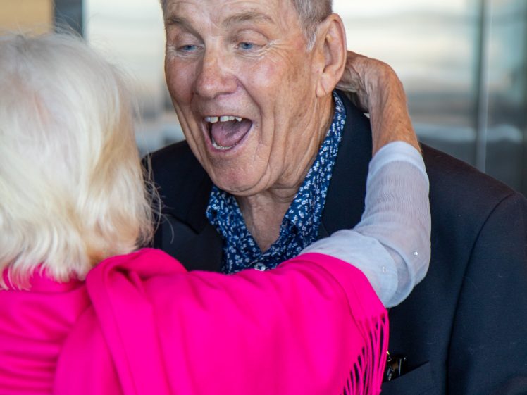 Image of An elderly man with grey hair and a blue patterned shirt smiles widely as he is embraced by a woman with white hair wearing a bright pink shawl.