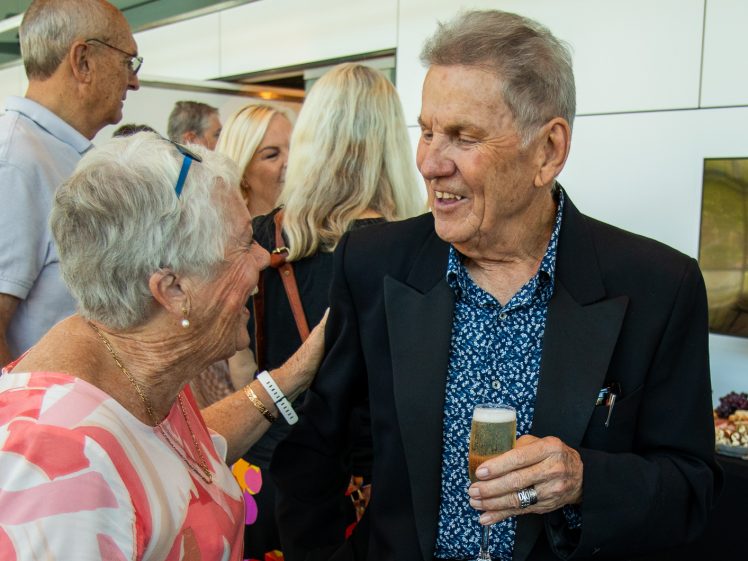 Image of An older man in a dark suit holds a drink and smiles whilst talking to an older woman in glasses and a pink-patterned top at a social gathering, with other people chatting in the background.