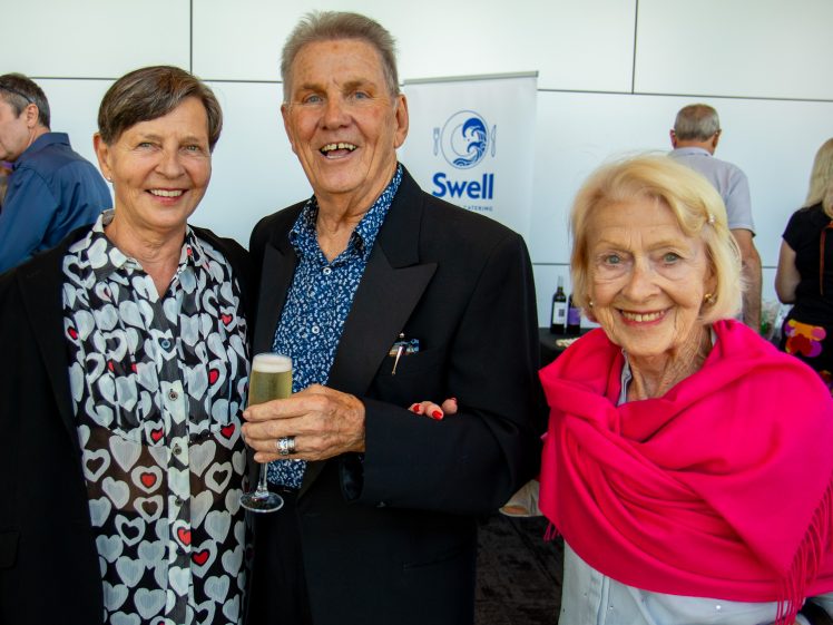 Image of Three smiling older adults pose together at an indoor event, with one man holding a drink. Two women, one wearing a heart-patterned shirt and the other a bright pink shawl, stand beside him. Other people and a Swell sign are in the background.