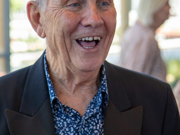Image of An older man with grey hair smiles while holding a drink. He is wearing a dark blazer over a blue patterned shirt. The background is softly blurred with another person visible.