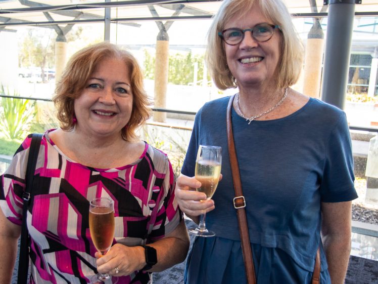 Image of Two smiling women stand indoors, each holding a glass of champagne. One wears a pink patterned dress and the other a blue dress with glasses and a brown cross-body bag. Sunlight streams through large windows behind them.