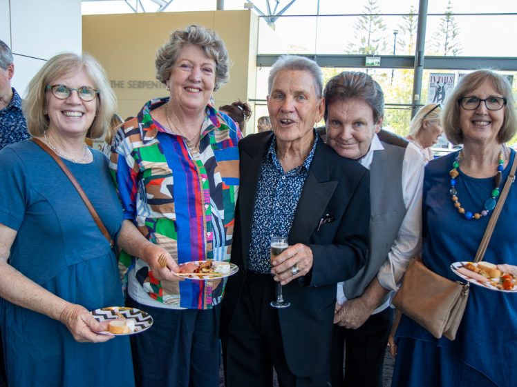 Image of Five adults stand together at a social event, smiling at the camera. Four hold plates of food; one holds a glass of champagne. They stand indoors near large windows with trees visible outside.