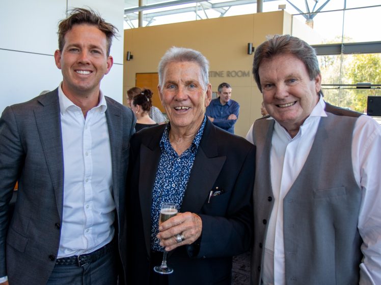 Image of Three men in formal attire stand together indoors, smiling at the camera. The man in the centre holds a glass of champagne. Sunlight streams in through large windows in the background.