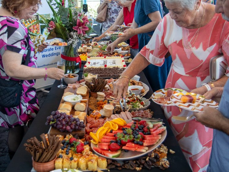 Image of A group of people serve themselves food from a buffet table filled with desserts, fruit, cheese, and snacks at an indoor event. Plates, cutlery, and a flower arrangement are also visible on the table.