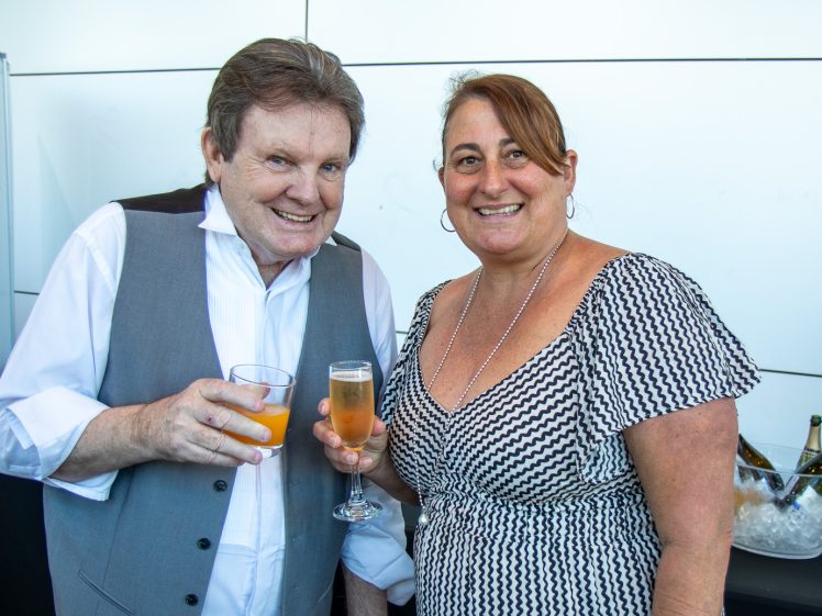 Image of A man and woman smiling at the camera, holding drinks. The man wears a white shirt and grey waistcoat; the woman wears a black and white patterned dress. Bottles and ice are visible in the background.