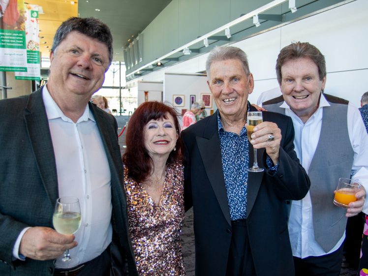 Image of Four adults, three men in suits and a woman in a sparkly dress, smile and hold drinks while posing together at a social event in a bright indoor setting.