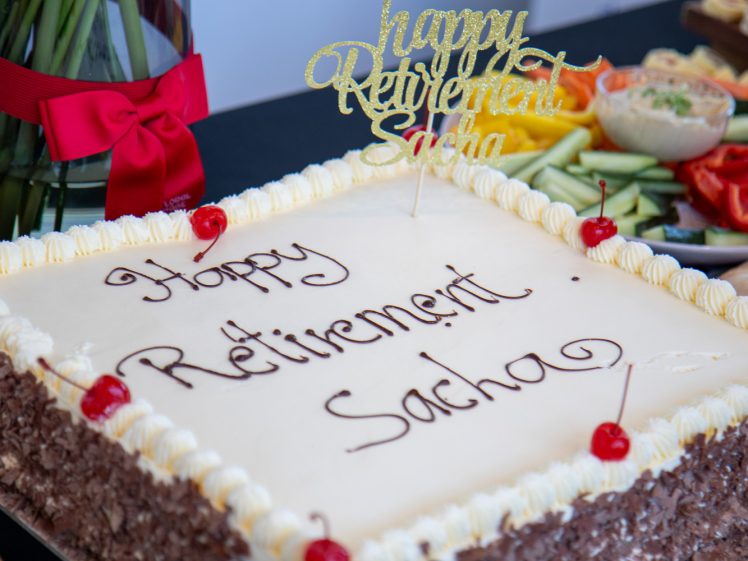 Image of A rectangular cake with white icing, chocolate shavings on the sides, cherries on top, and “Happy Retirement Sacha” written in chocolate. A gold cake topper and a platter of vegetables are visible in the background.