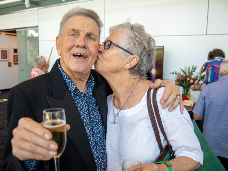 Image of An older woman kisses an older man on the cheek as he smiles, holding a glass of champagne at a social event. They stand close together in a bright, modern room with people and art in the background.