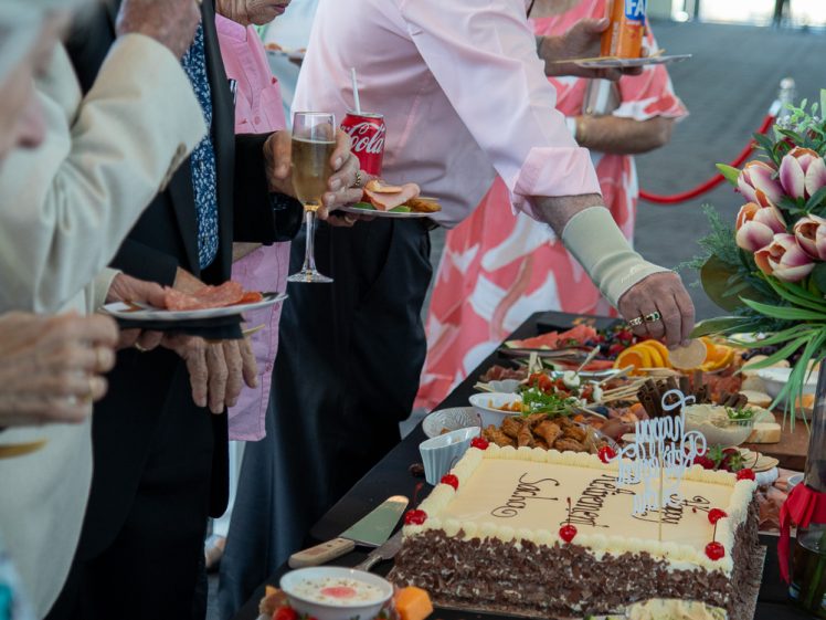Image of A group of elderly people stand around a table filled with food, desserts, and a large decorated cake. Some are serving themselves, and one person holds a glass of sparkling wine. The atmosphere appears celebratory.