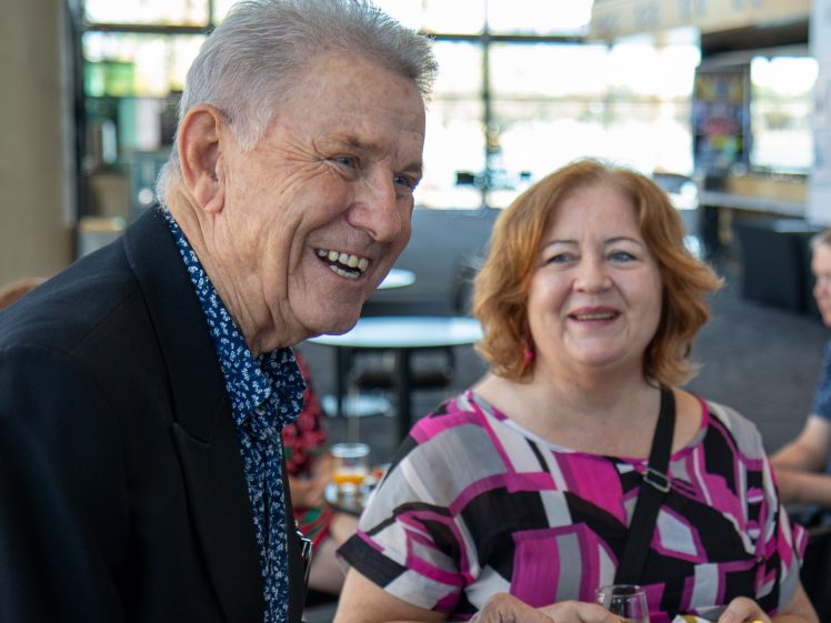 Image of An older man in a dark jacket and a smiling woman in a pink patterned top enjoy drinks and snacks together indoors, with bright windows and other people in the background.