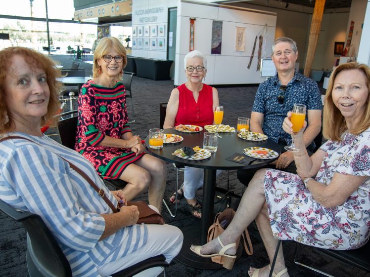 Image of Five smiling adults sit around a table with drinks and plates of food in a modern art gallery, enjoying a social gathering. Colourful artwork is displayed on the walls behind them.