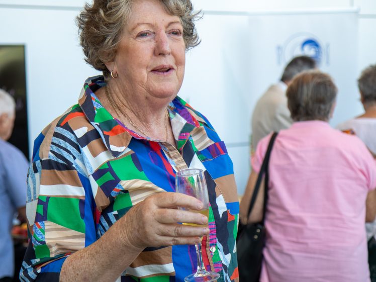 Image of An older woman with curly grey hair holds a drink in a glass and smiles. She wears a colourful patterned shirt. Several people are gathered and talking in the background at what appears to be a social event.