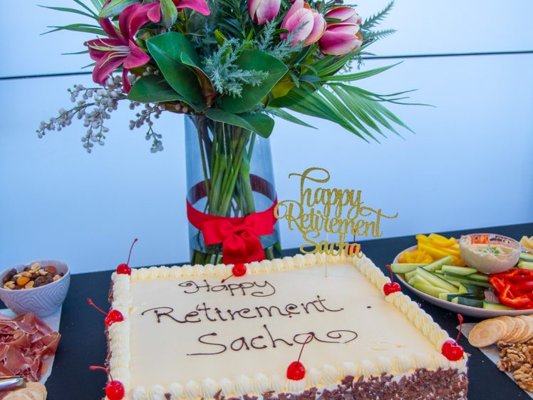 Image of A rectangular cake with Happy Retirement Sacha written on it sits on a table beside a bouquet of pink flowers, fruit, nuts, crackers, and a “Happy Retirement” cake topper. A cake knife and server are in front.