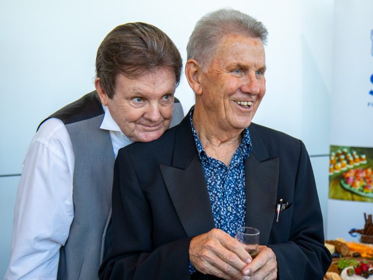 Image of Two elderly men dressed in formal attire stand close together, smiling. One man leans his head on the other’s shoulder, holding a drink, with food platters visible in the background.