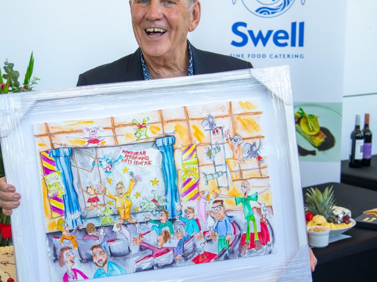 Image of An older man smiles whilst holding a colourful framed cartoon-style artwork. Behind him is a Swell Fine Food Catering banner and a table with food, wine bottles, and flowers.