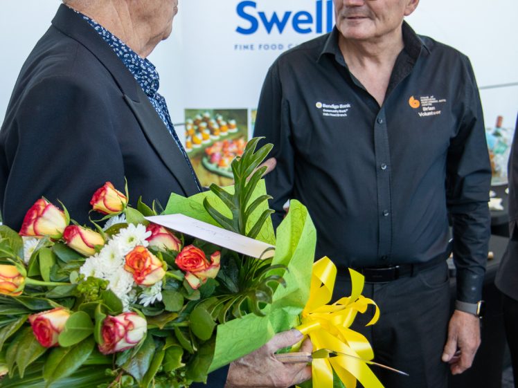 Image of Two older men stand facing each other indoors; one holds a bouquet of flowers with yellow ribbon. Behind them is a sign reading “Swell Fine Food.” Food platters are visible on a table in the background.