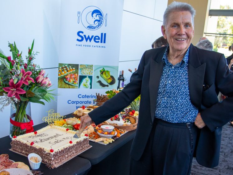 Image of An older man in a suit smiles whilst cutting a decorated cake at a catered event. The table displays flowers and various foods. A Swell Fine Food Catering banner stands in the background.