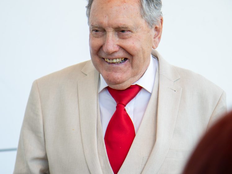 Image of An older man in a light beige suit, white shirt, and bright red tie is smiling warmly. He holds a glass in his hand, and the background is blurred and white.