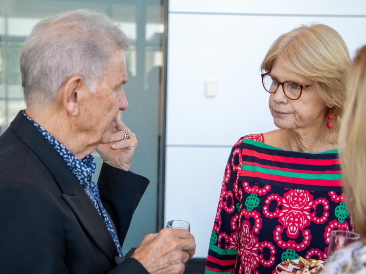 Image of An older man in a suit talks to an older woman wearing glasses and a colourful patterned top. She holds a plate of food, listening attentively. Another person’s shoulder is partially visible in the foreground.