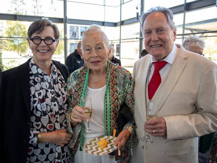 Image of Three older adults dressed formally stand together indoors, smiling at the camera. The woman in the centre holds a glass of champagne and a plate of snacks. Other people are visible in the background.